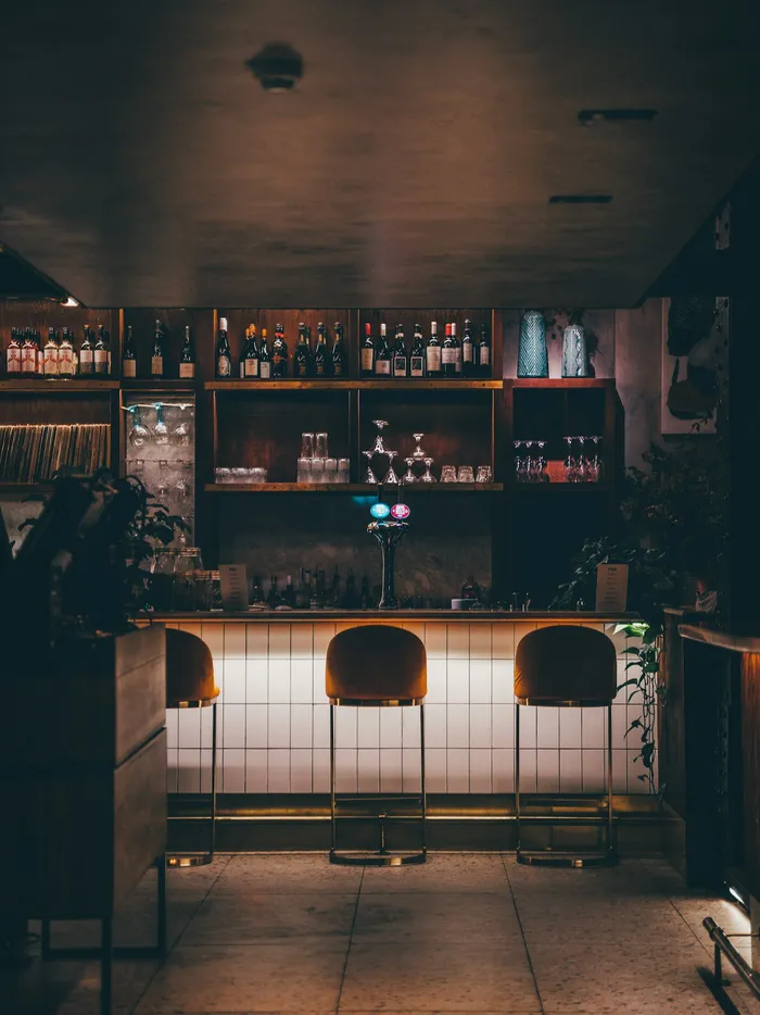 Comptoir de bar en bois et tabourets velours, étagères de bouteilles en lumière tamisée